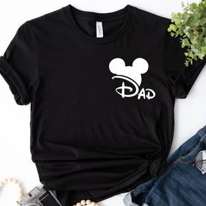 K&ouml;nnte beinhalten: Schwarzes T-Shirt mit einem wei&szlig;en Mickey-Maus-Kopf und dem Wort "DAD" in einer Disney-Schrift.