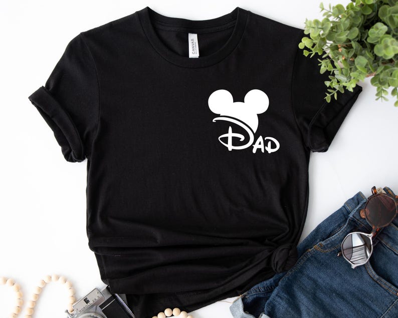 K&ouml;nnte beinhalten: Schwarzes T-Shirt mit einem wei&szlig;en Mickey-Maus-Kopf und dem Wort "DAD" in einer Disney-Schrift.