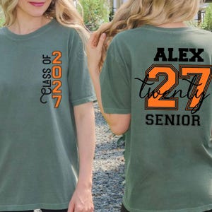 Może przedstawiać: Szałwiowo-zielony t-shirt z pionową pomarańczowo-czarną grafiką na piersi z napisem "CLASS OF 2027". Z tyłu koszulki znajduje się imię "ALEX" w kolorze czarnym, duża pomarańczowa liczba "27" z czarnym obrysem oraz słowo "SENIOR".