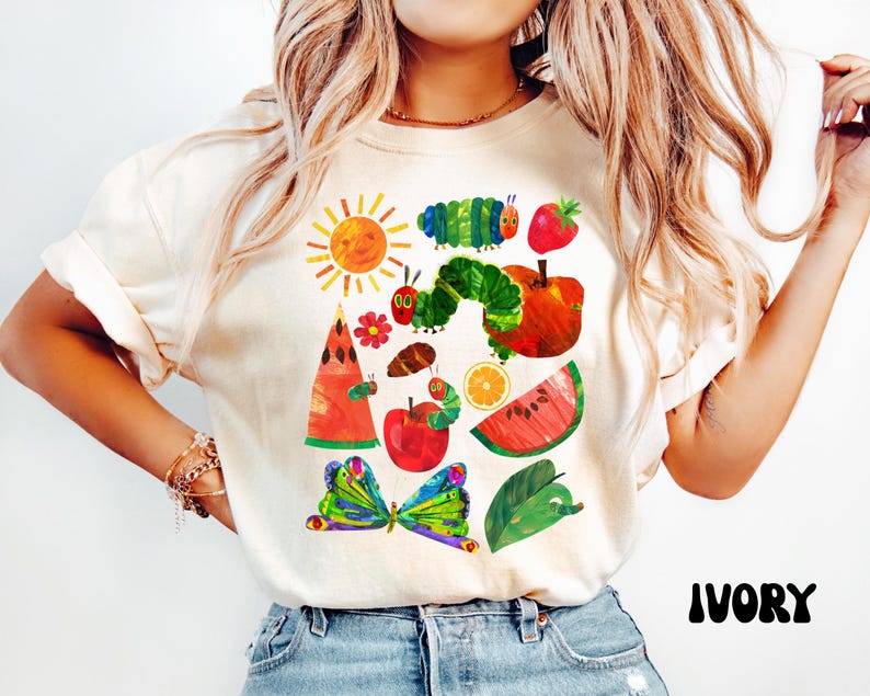Puede incluir: Camiseta de color marfil con una colorida ilustraci&oacute;n de una oruga, un sol, frutas y una mariposa. El dise&ntilde;o incluye una oruga verde, una manzana roja y una rodaja de sand&iacute;a. La palabra "IVORY" est&aacute; impresa en negro en la parte inferior.