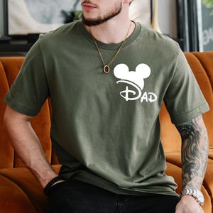 K&ouml;nnte beinhalten: Ein gr&uuml;nes T-Shirt mit einem wei&szlig;en Mickey-Maus-Kopf und dem Wort "DAD" darunter. Der Mickey-Maus-Kopf tr&auml;gt einen wei&szlig;en Hut mit einer wei&szlig;en Schleife.