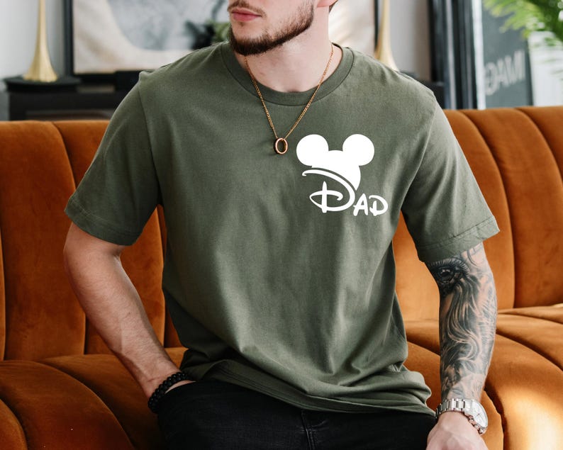 K&ouml;nnte beinhalten: Ein gr&uuml;nes T-Shirt mit einem wei&szlig;en Mickey-Maus-Kopf und dem Wort "DAD" darunter. Der Mickey-Maus-Kopf tr&auml;gt einen wei&szlig;en Hut mit einer wei&szlig;en Schleife.