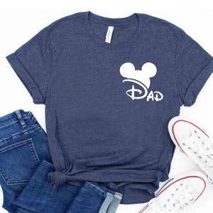 K&ouml;nnte beinhalten: Ein marineblaues T-Shirt mit einem wei&szlig;en Mickey-Maus-Kopf und dem Wort "DAD" darunter. Das Shirt ist gefaltet und auf einer wei&szlig;en Oberfl&auml;che abgelegt.
