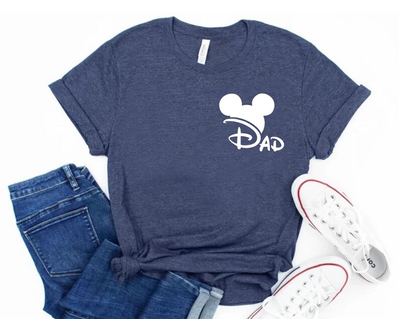 K&ouml;nnte beinhalten: Ein marineblaues T-Shirt mit einem wei&szlig;en Mickey-Maus-Kopf und dem Wort "DAD" darunter. Das Shirt ist gefaltet und auf einer wei&szlig;en Oberfl&auml;che abgelegt.
