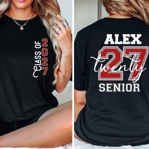 Może przedstawiać: Czarny t-shirt z nadrukiem z przodu o treści CLASS OF 2027 w kolorze czerwonym i białym. Z tyłu koszulki znajduje się imię ALEX nad dużą czerwoną liczbą 27, z napisem twenty białą kursywą oraz słowem SENIOR wydrukowanym poniżej.