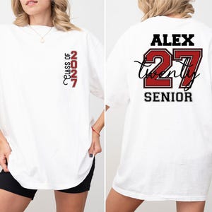 Może przedstawiać: Biały t-shirt z pionową czerwono-czarną grafiką na lewej piersi z napisem Class of 2027. Z tyłu znajduje się imię Alex w kolorze czarnym, numer 27 w kolorze czerwonym z czarnym obrysem oraz słowo Senior napisane czarną czcionką.