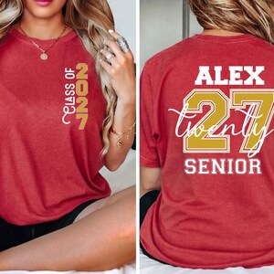 Może przedstawiać: Czerwony t-shirt z kr&oacute;tkim rękawem z napisem "CLASS OF 2027" w kolorze złotym na lewej piersi. Z tyłu znajduje się imię "ALEX" na biało, liczba "27" na złoto, słowo "twenty" białą kursywą oraz napis "SENIOR" w kolorze białym.
