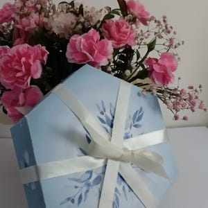 Puede incluir: Una caja de regalo hexagonal azul claro atada con una cinta blanca, decorada con un diseño floral. Una cesta de mimbre llena de claveles rosas y paniculata en el fondo, creando una estética suave y romántica.