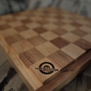 Könnte beinhalten: Ein Schachbrett aus Holz mit einem hellen und dunklen Holzmuster. Das Brett besteht aus zwei verschiedenen Holzarten, die einen Kontrasteffekt erzeugen. Das Brett hat ein Logo in der unteren rechten Ecke, das "The Deep South Woodshop Est. 2021" lautet.