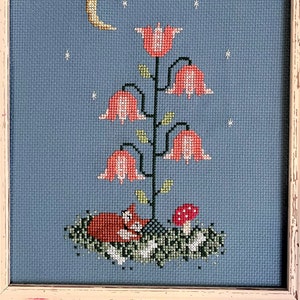 Peut inclure: Une broderie au point de croix représentant un renard roux dormant sous un croissant de lune et des étoiles, niché dans un lit de feuilles vertes et de fleurs blanches. Une plante haute avec des fleurs en forme de cloche rose pousse en arrière-plan.