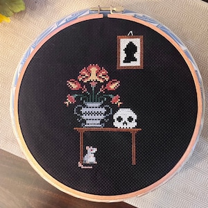 May include: A black and white cross-stitch embroidery of a still life with a vase of flowers, a skull, a mouse, and a framed silhouette. The vase is decorated with a white and blue pattern. The table is brown and the background is black.