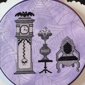 May include: A purple embroidery hoop with a cross-stitch design of a clock, a vase, and a chair. The design is in black and white and features a spiderweb pattern in the background.