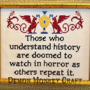 May include: A framed cross-stitch with a quote that reads "Those who understand history are doomed to watch in horror as others repeat it." The quote is framed with a red and yellow design.