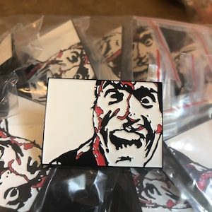 May include: A white enamel pin featuring a black and red illustration of a man's face with blood splatters. The man is screaming with his mouth open and his eyes wide.