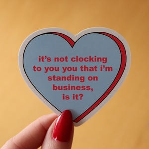 May include: A heart-shaped sticker with a light blue center and a red border. The sticker has the text "it's not clocking to you you that I'm standing on business, is it?" in red. The sticker is held by a hand with red nail polish.