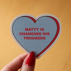 May include: A heart-shaped sticker with a light blue center and a red border. The sticker reads "MATTY IS CHANGING HIS TROUSERS" in red text. The sticker is held against a yellow background.