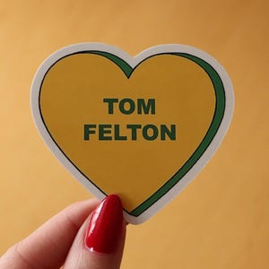 May include: A heart-shaped sticker with a yellow center and green border. The sticker has the name "TOM FELTON" printed in dark green. The sticker is held by a hand with red nail polish against a yellow background.
