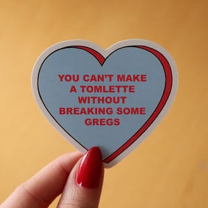 May include: A heart-shaped sticker with a light blue center and a red border. The sticker has the text "YOU CAN'T MAKE A TOMLETTE WITHOUT BREAKING SOME GREGS" in red. The sticker is held by a hand with red nail polish.