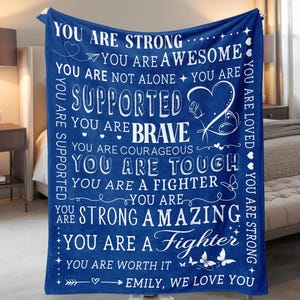 May include: A blue fleece blanket with white text. The blanket has inspirational phrases such as "You are strong", "You are brave", and "You are loved". The blanket also includes the text "Emily, we love you".