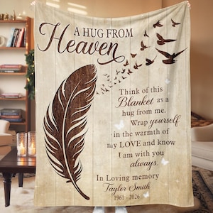 May include: A beige blanket with a wood-grain design, featuring a large brown feather and flying birds. Text includes "A HUG FROM Heaven" and a message of comfort and remembrance. The blanket is a cozy home decor item.