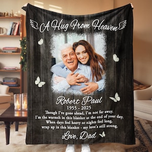 May include: Black blanket with a wooden plank design, featuring a photo of a man and woman embracing. Text includes "A Hug From Heaven," "Robert Paul 1955-2025," and a heartfelt message. White butterflies accent the design.