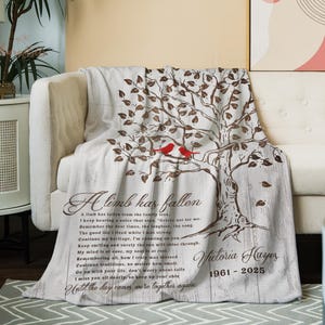 May include: A light-colored throw blanket draped over a cream-colored sofa. The blanket features a tree design with red birds and text that reads "A limb has fallen." The blanket is a cozy home decor item.