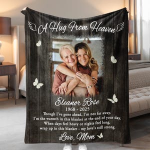 May include: Black blanket with a wooden plank design, featuring a photo of two women embracing. Text includes "A Hug From Heaven," "Eleanor Rose 1968-2025," and a loving message. White butterflies are scattered across the blanket.