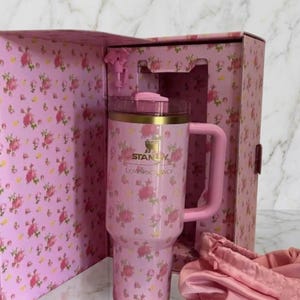 May include: A pink Stanley tumbler with a floral pattern, featuring a gold rim, a pink lid, and a matching handle. The tumbler is housed in a matching pink floral box with the text Stanley and LoveShackFancy printed on the front of the cup.