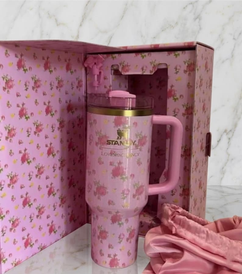 May include: A pink Stanley tumbler with a floral pattern, featuring a gold rim, a pink lid, and a matching handle. The tumbler is housed in a matching pink floral box with the text Stanley and LoveShackFancy printed on the front of the cup.