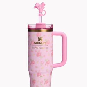 May include: A light pink insulated tumbler with a floral pattern, featuring a gold Stanley logo and the text LOVESHACKFANCY. The tumbler has a matching pink handle, a clear lid, and a straw topper shaped like a pink bow on a clear straw.