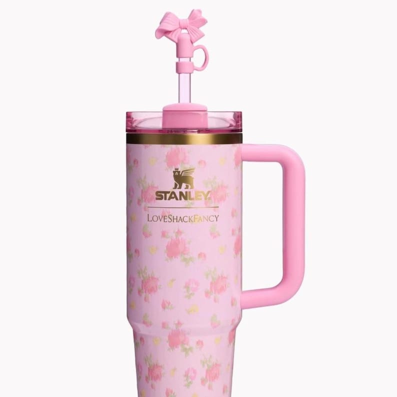 May include: A light pink insulated tumbler with a floral pattern, featuring a gold Stanley logo and the text LOVESHACKFANCY. The tumbler has a matching pink handle, a clear lid, and a straw topper shaped like a pink bow on a clear straw.