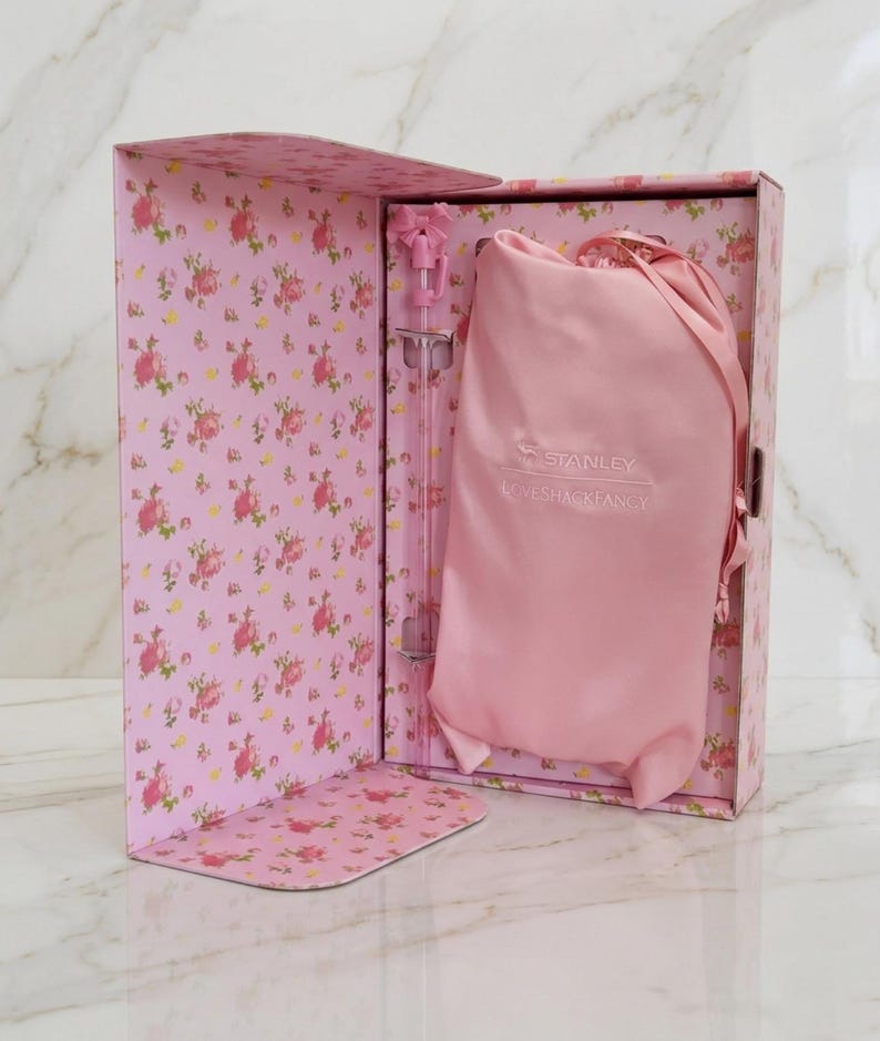 May include: A pink floral patterned box containing a light pink drawstring pouch with the Stanley and LoveShackFancy logos printed in white, alongside a clear straw featuring a small pink bow detail at the top, presented in a gift set.