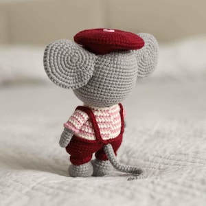 May include: A handmade crochet mouse doll, viewed from the back, wearing a burgundy beret, overalls, and a pink and white striped shirt. The mouse has large gray ears and a long gray tail. The doll is made of yarn.