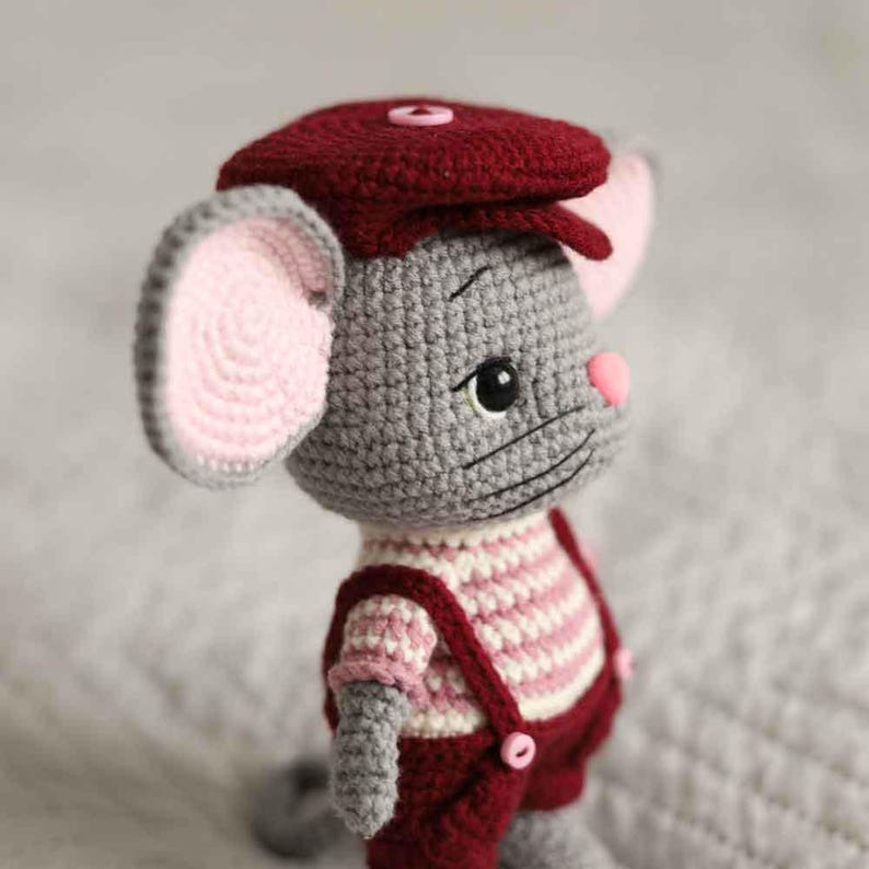 May include: Crocheted mouse doll wearing a burgundy cap and overalls. The mouse has gray fur, pink ears and nose, and a striped shirt. The doll is handmade and has button details.