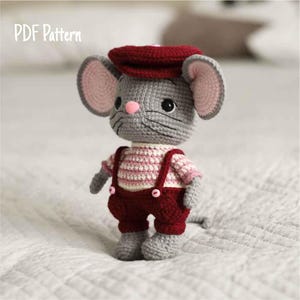 Może przedstawiać: Ręcznie robiona szydełkowa lalka myszy w czerwonej czapce, czerwonych ogrodniczkach z różowymi guzikami i w paski. Mysz ma szare futro, różowe uszy, różowy nos i czarne oczy. Widoczny jest tekst "PDF Pattern".