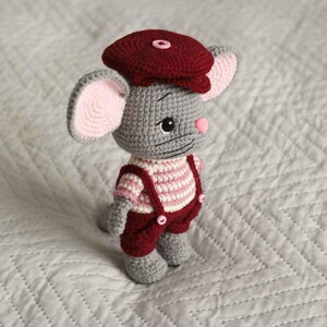 May include: A crocheted gray mouse wearing a burgundy cap and overalls. The mouse has pink ears and nose, and a striped pink and white shirt. The overalls have burgundy buttons. The toy is handmade and sits on a white quilted surface.