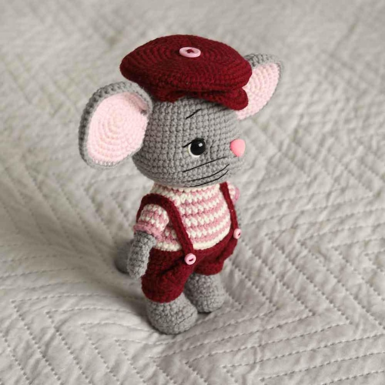 May include: A crocheted gray mouse wearing a burgundy cap and overalls. The mouse has pink ears and nose, and a striped pink and white shirt. The overalls have burgundy buttons. The toy is handmade and sits on a white quilted surface.