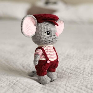 May include: A crocheted mouse doll wearing a burgundy newsboy cap, overalls, and a pink and white striped shirt. The mouse has gray fur, pink ears, and a pink nose. The doll is standing on a white surface.