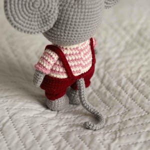 May include: A handmade crochet mouse toy, viewed from the back, wearing a red and white checkered shirt and red overalls. The mouse is gray with large ears and a long tail. The toy is sitting on a white, quilted surface.