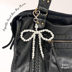 May include: A white pearl bow bag charm with a silver clasp, attached to a black leather handbag. The charm has a bow design with pearl strands. The text "Coquette Pearl Bow Bag Charm" is visible.