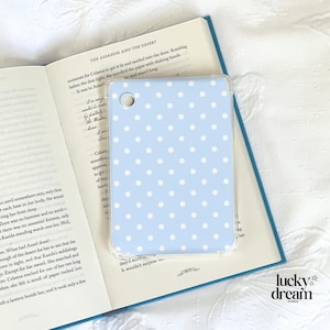 May include: A light blue phone case with a white polka dot design, placed on an open book. The book's pages show text from "The Assassin and the Desert". The case has a clear border and the Lucky Dream Studios logo is visible.