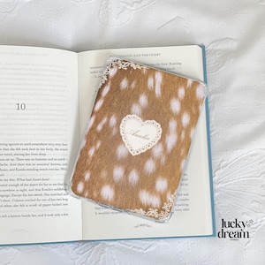 May include: A phone case with a brown and white spotted pattern, resembling deer fur, is placed on an open book. The case has a white heart-shaped design with the name "Amelia" and a decorative lace border.