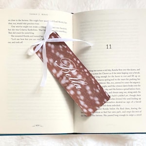 May include: A decorative bookmark with a white ribbon, featuring a brown and white spotted pattern. The word "Bella" is written in elegant script. The bookmark is placed in an open book.