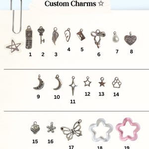 May include: A collection of nineteen numbered metal and resin charms labeled Custom Charms. The set includes shapes like a flip phone, key, wing, envelope, bow, pearl, heart, moon, star, paw print, strawberry, flower, butterfly, and star-shaped rings.