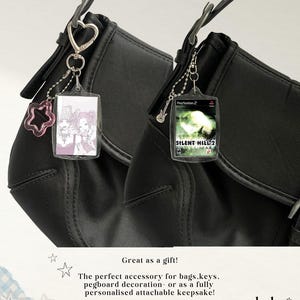 May include: Two black shoulder bags featuring rectangular clear plastic keychains. One keychain displays a manga-style illustration of two characters, while the other shows the Silent Hill 2 PlayStation 2 game cover. Both have metal hardware.