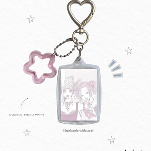 May include: A rectangular clear acrylic keychain featuring a double-sided print of two illustrated figures in pink tones. The keychain includes a silver heart-shaped clasp and a translucent pink star-shaped charm attached by a small metal ball chain.