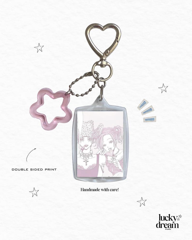 May include: A rectangular clear acrylic keychain featuring a double-sided print of two illustrated figures in pink tones. The keychain includes a silver heart-shaped clasp and a translucent pink star-shaped charm attached by a small metal ball chain.