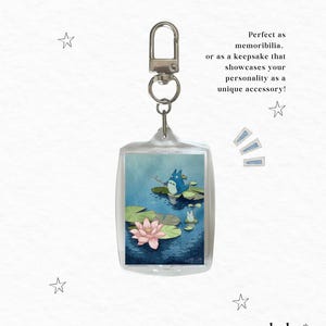 May include: A rectangular clear acrylic keychain featuring an illustration of a blue creature fishing on a lily pad in a pond with a pink lotus flower. The text reads: Perfect as memorabilia. or as a keepsake that showcases your personality as a unique accessory!