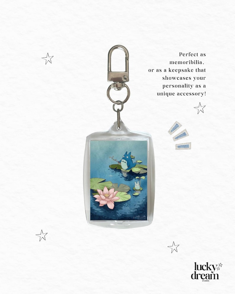 May include: A rectangular clear acrylic keychain featuring an illustration of a blue creature fishing on a lily pad in a pond with a pink lotus flower. The text reads: Perfect as memorabilia. or as a keepsake that showcases your personality as a unique accessory!