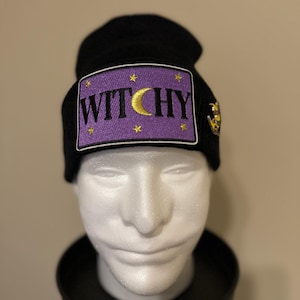 May include: Black beanie with a purple patch featuring the word "WITCHY" in white letters, a gold crescent moon, and gold stars. A gold emblem is on the side. The beanie is displayed on a white mannequin head.